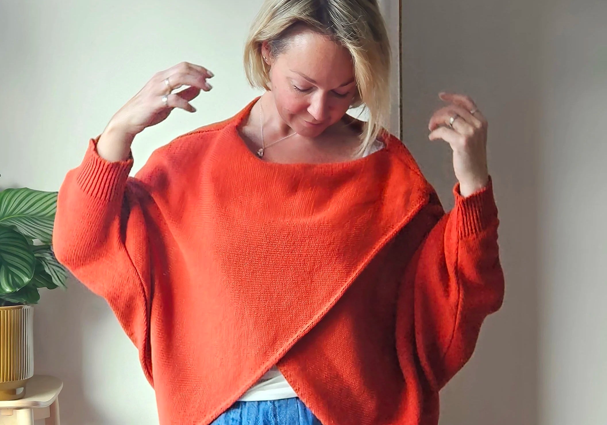 Woman wearing an orange sweater with a plant and white wall in the background