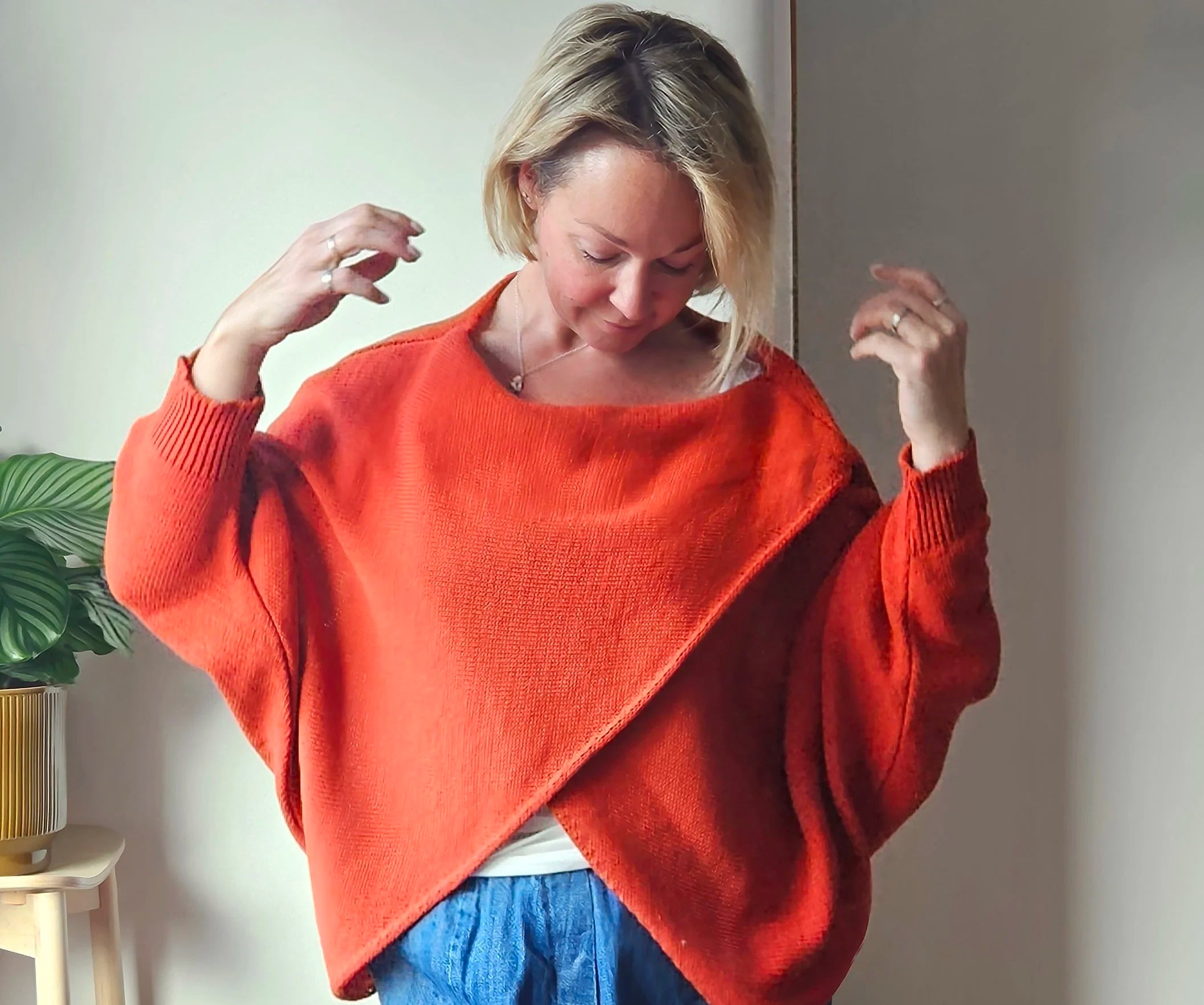 Woman wearing an orange sweater with a plant and white wall in the background