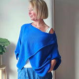 Woman wearing a blue cross front jumper standing against a plain wall with a plant to the side.