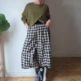 Person wearing a green sweater, black and white checkered pants, and black high-top sneakers in a room with a plant and wooden floor.