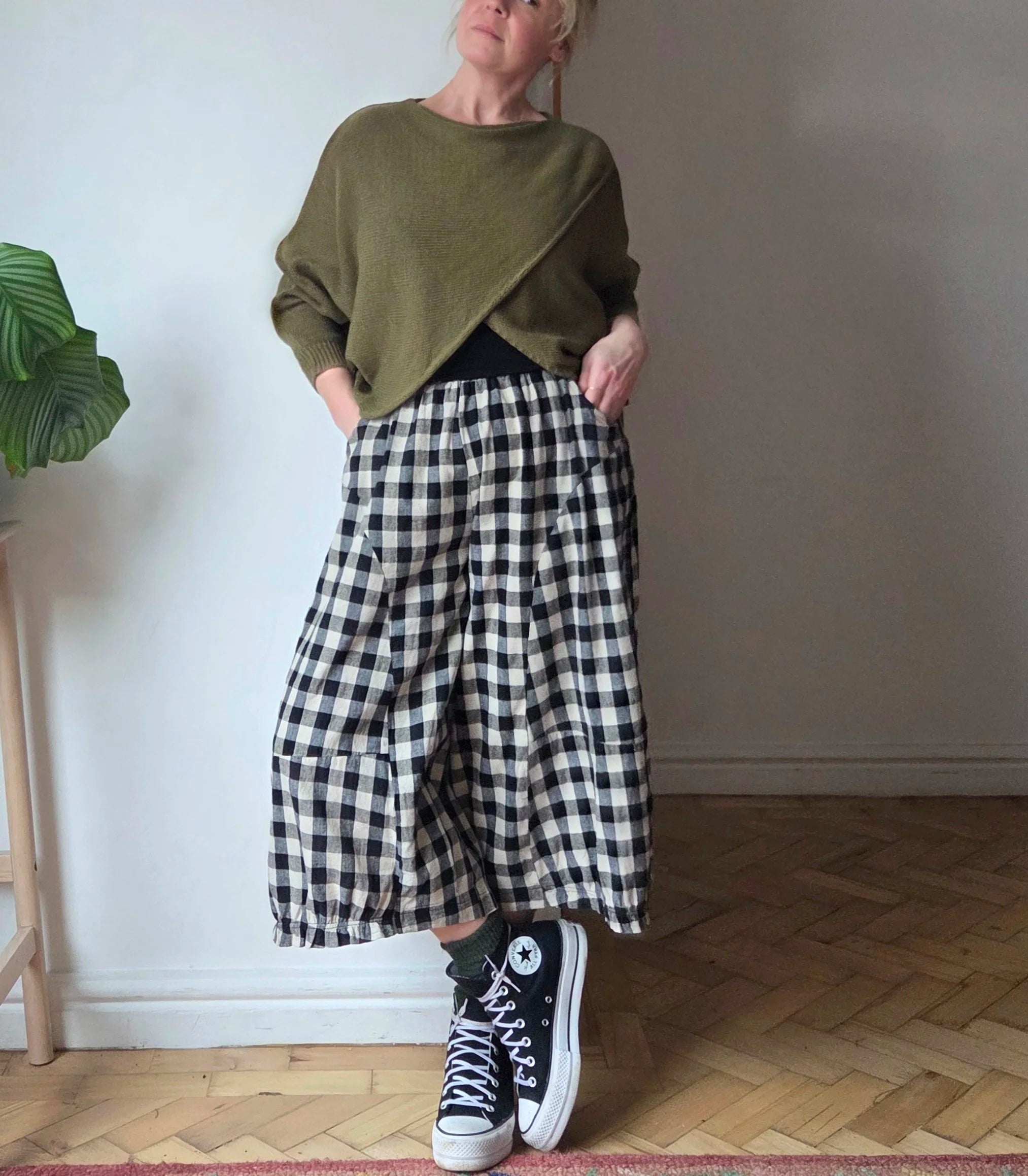 Person wearing a green sweater, black and white checkered pants, and black high-top sneakers in a room with a plant and wooden floor.