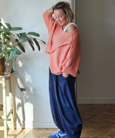 Person wearing a pink oversized sweatshirt and blue pants standing indoors with a plant in the background.