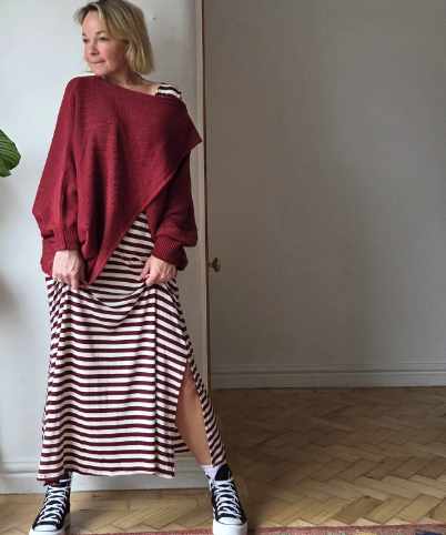Woman wearing a red sweater and striped skirt standing in a room with wooden flooring.