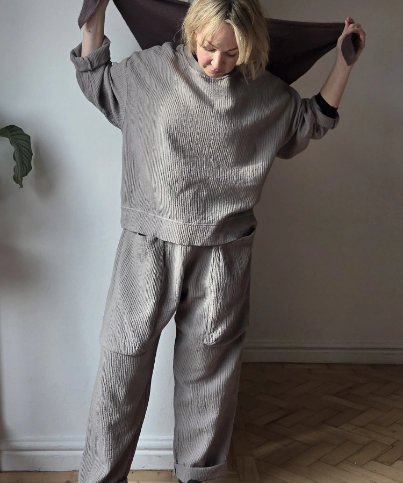 Person wearing a gray knit outfit holding a brown fabric against a plain wall.